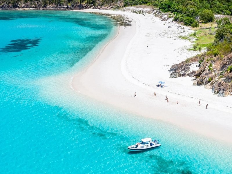A white sand spit curling into turquoise water below a wooded headland, a small boat at anchor off the point
